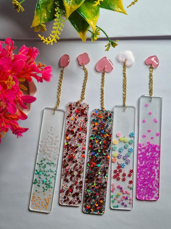 Five clear resin bookmarks filled with coloured glitter, each with a gold chain and charm